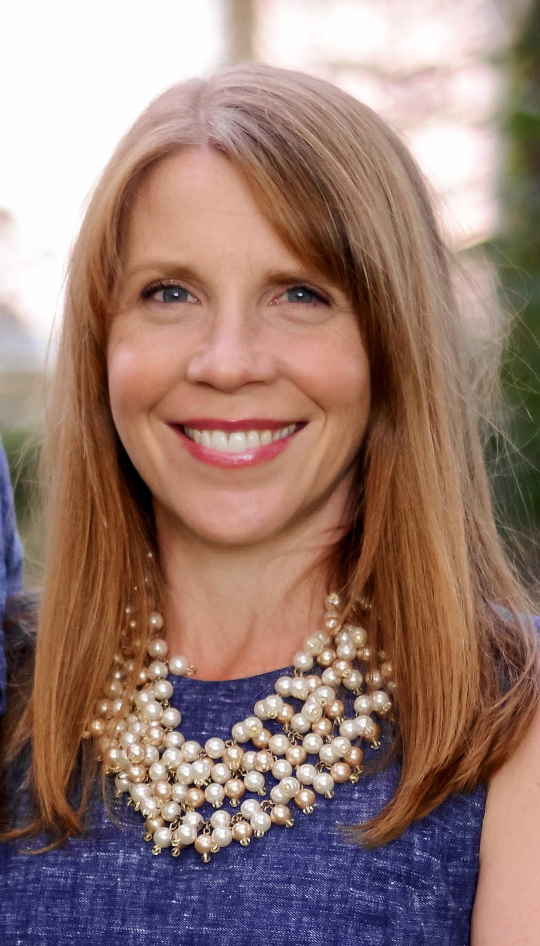 Woman smiling in a professional headshot, wearing a blue top and statement pearl necklace outdoors